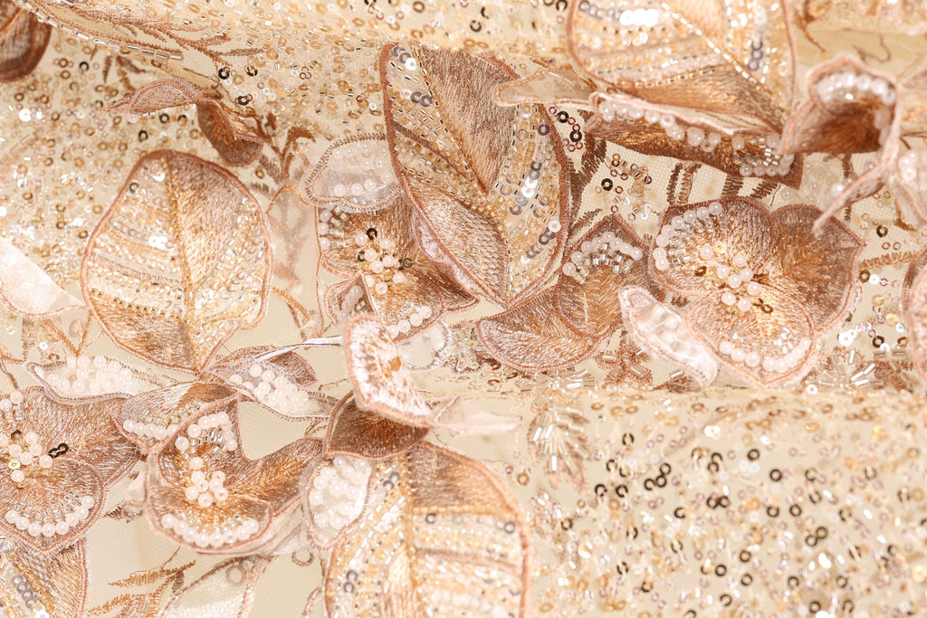 The image is a close-up of a fabric with intricate gold and white embroidered designs, including flowers and leaves. The fabric appears to be a delicate and ornate material, possibly used for clothing or other decorative purposes.