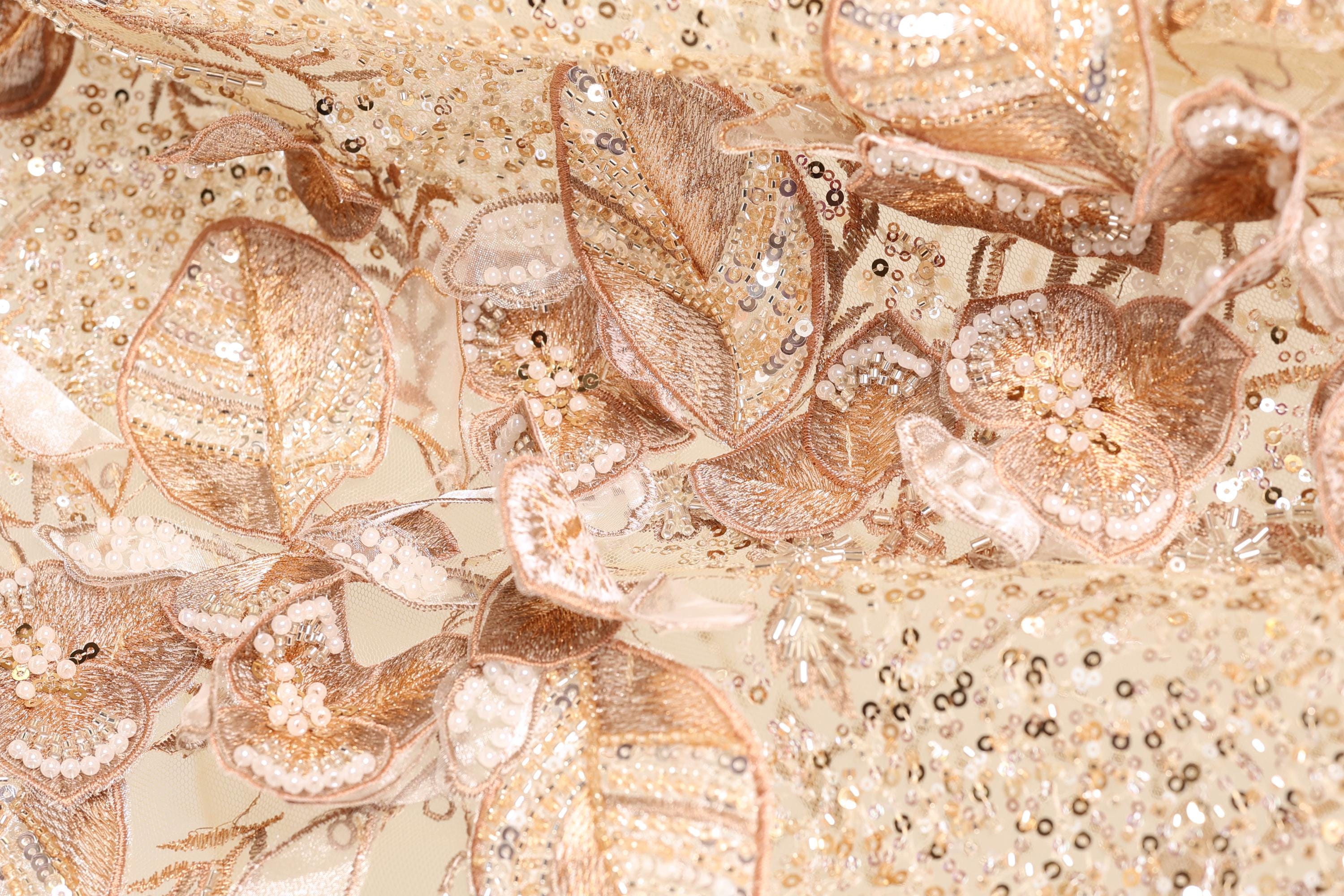 The image is a close-up of a fabric with intricate gold and white embroidered designs, including flowers and leaves. The fabric appears to be a delicate and ornate material, possibly used for clothing or other decorative purposes.