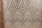 a close-up view of a fabric with a repeating pattern of diamond shapes and small dots in shades of brown and beige. The pattern is intricate and detailed, with the diamonds and dots arranged in a zigzag or diagonal manner. The fabric appears to be made of a shiny, sequined material, which adds to the visual appeal of the design.