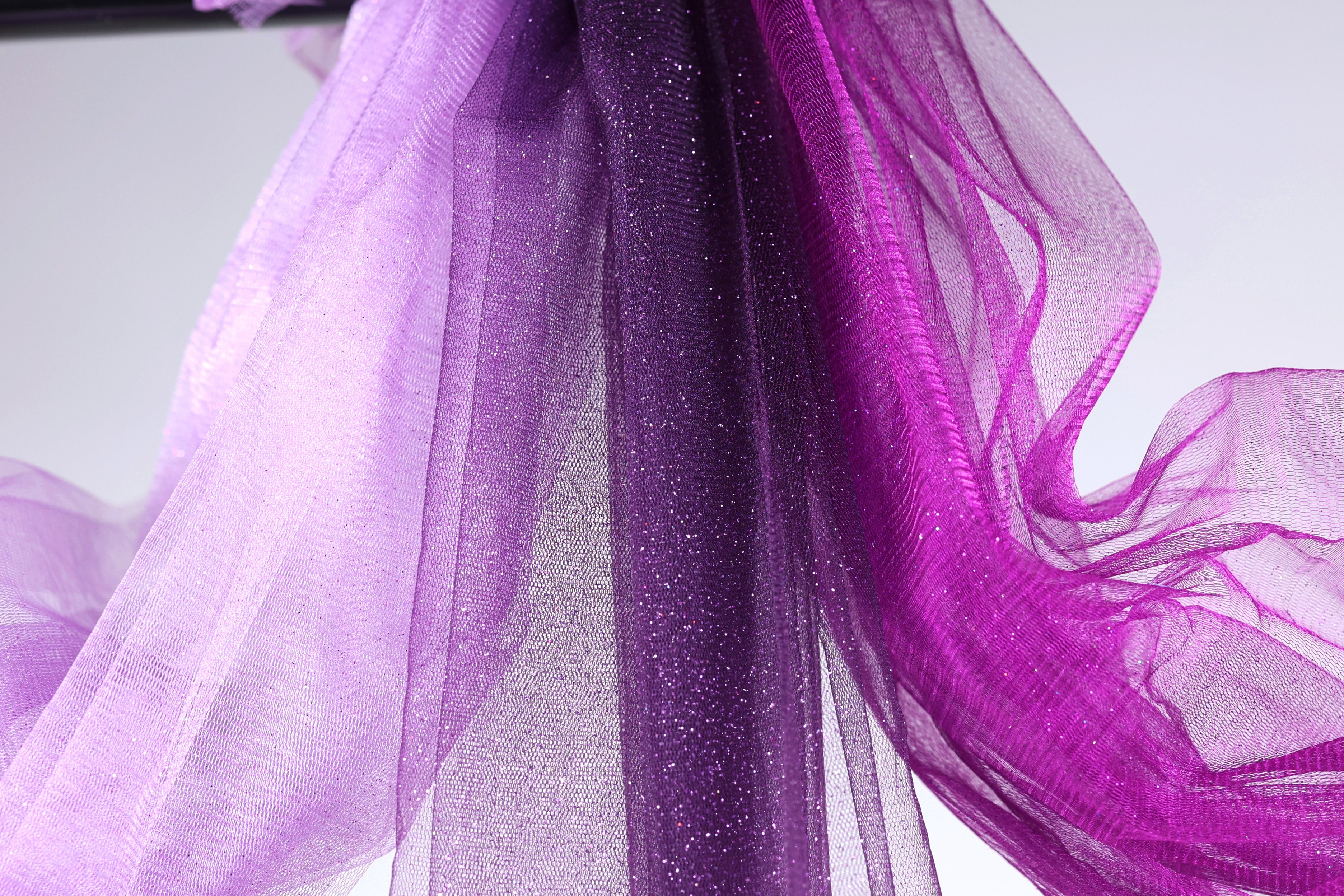 a close-up view of a piece of purple and white fabric, with the fabric appearing to be made of sheer material. The fabric has a shiny, glittery texture, adding a touch of sparkle to the overall appearance. The fabric is hanging in the air, creating a sense of movement and dynamism.