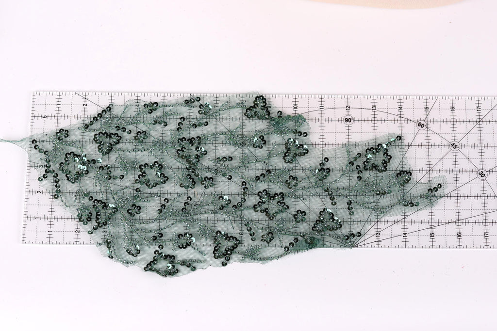 a transparent green fabric with numerous small black sequins or beads scattered across it, placed on a transparent ruler or measuring mat.