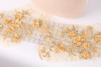 a close-up view of a delicate, ornate fabric with a floral pattern. The fabric is adorned with numerous small gold flowers, creating a visually striking and intricate design. The flowers are arranged in a way that they appear to be growing out of the fabric, adding a sense of depth and dimension to the piece. The fabric is spread out on a white surface, allowing the viewer to appreciate the intricate details and craftsmanship of the design.