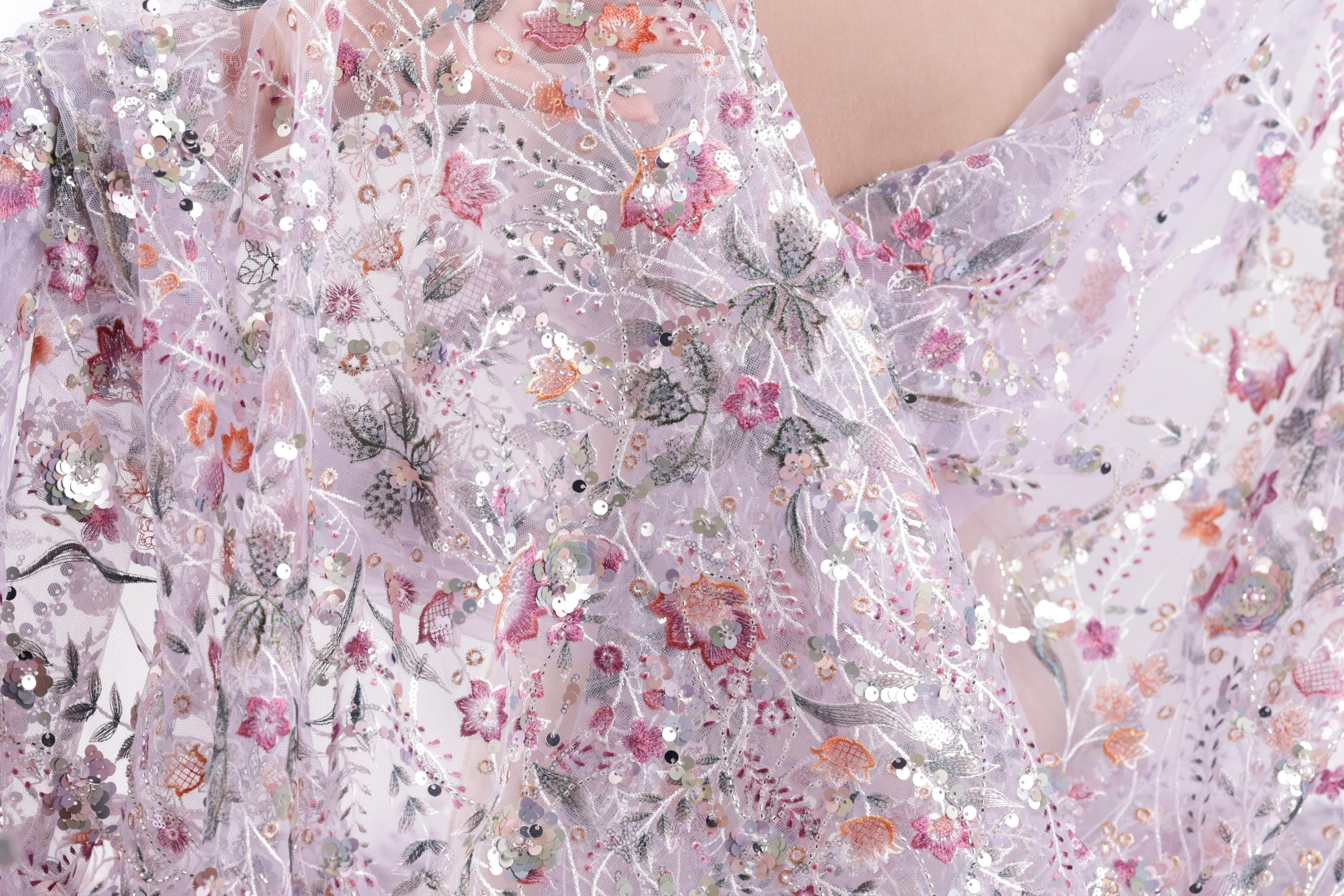a close-up view of a delicate, intricately embroidered fabric with a floral pattern. The fabric appears to be made of sheer material, giving it a soft and delicate appearance. The embroidery features a variety of flowers and leaves in shades of pink, purple, and green, creating a visually appealing and detailed design.
