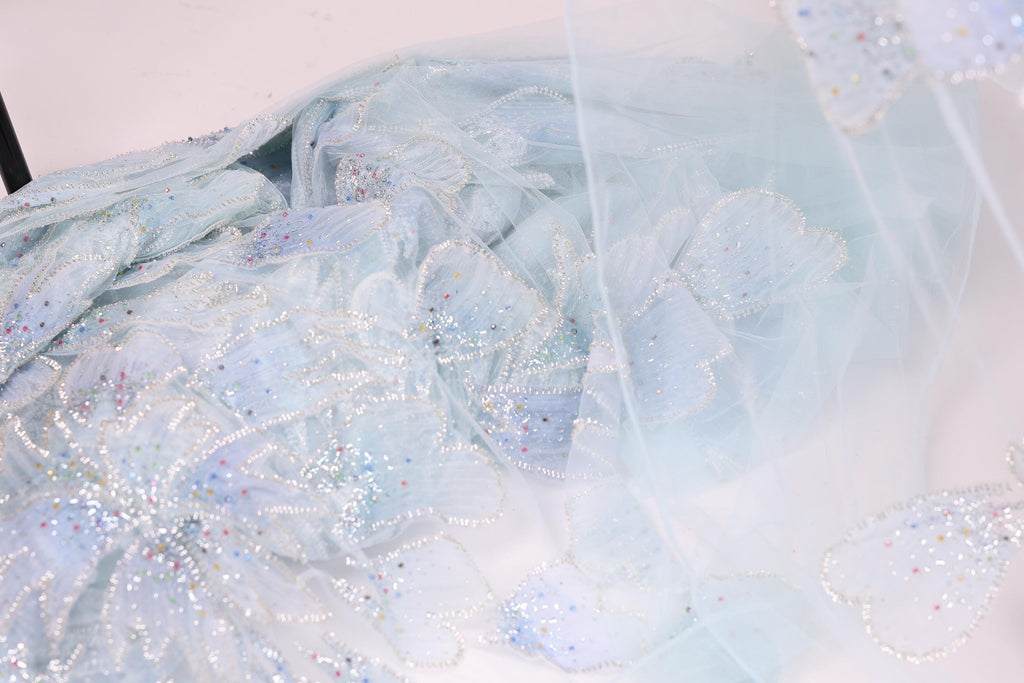 a close-up view of a delicate, light blue fabric with a floral pattern. The fabric appears to be made of sheer material, giving it a soft and airy appearance. The intricate design of the fabric, with its numerous small flowers or petals, creates a visually appealing and delicate texture.