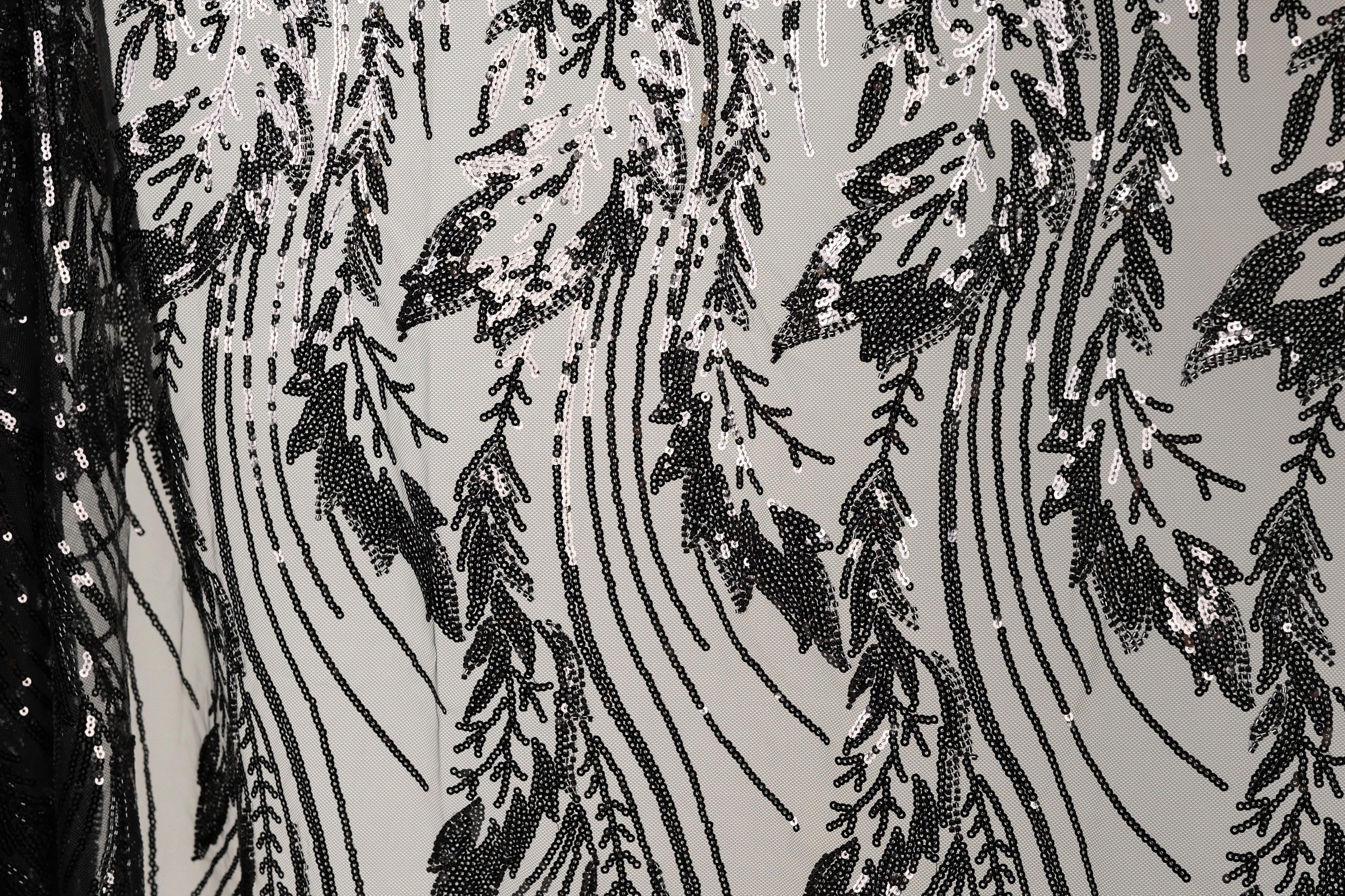 a close-up view of a black and white fabric with a repeating pattern of sequin designs. The pattern consists of various shapes and lines, creating a visually striking and intricate design. The fabric appears to be made of a material that resembles lace or sheer fabric, with the sequins adding a touch of sparkle and texture to the overall appearance.