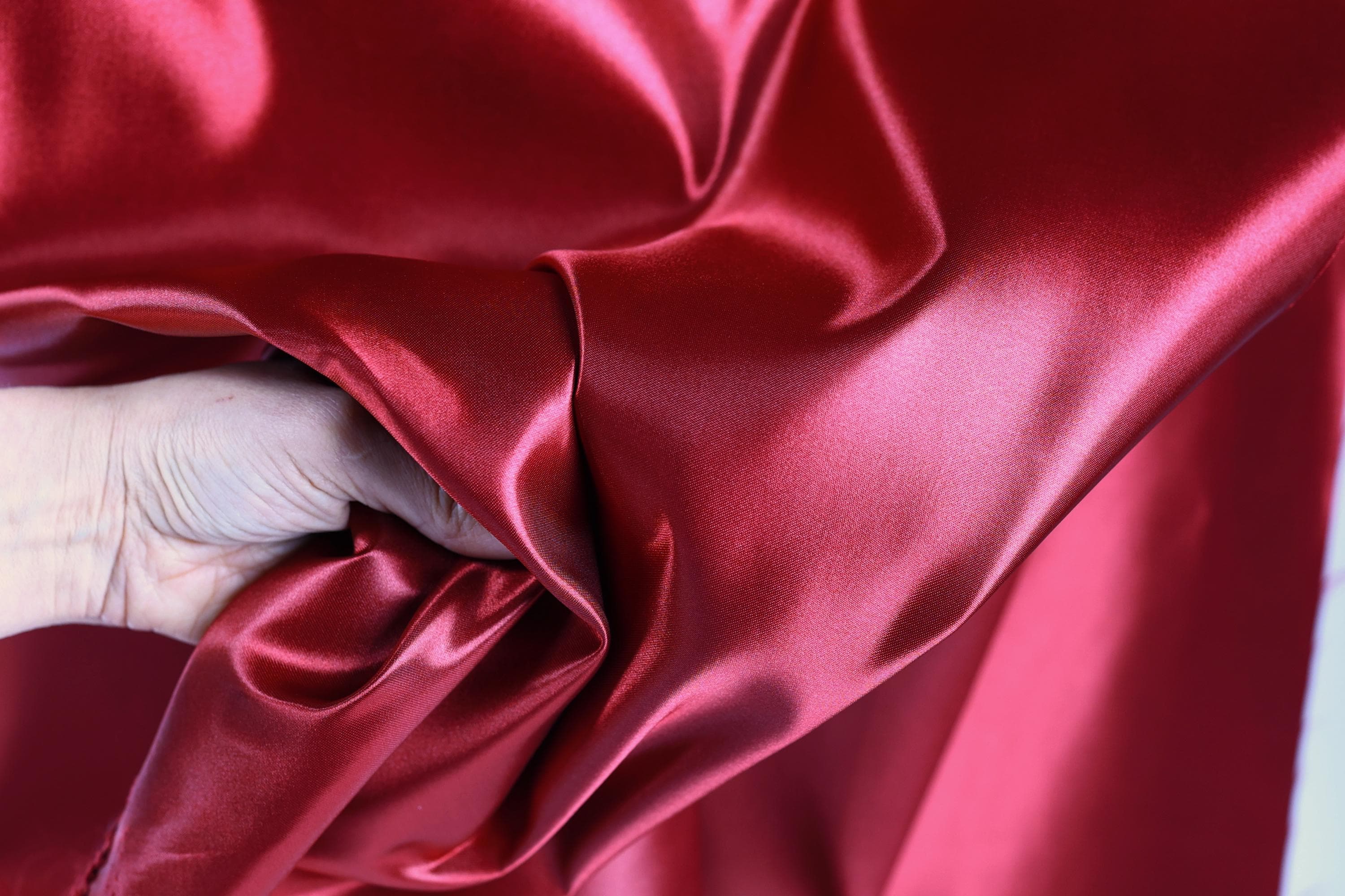 A hand is holding a piece of red satin fabric, which appears to be shiny and smooth.