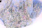 The image is a close-up of a delicate, intricately embroidered fabric with a floral pattern. The fabric appears to be made of a sheer, light-colored material, giving it a soft and delicate appearance. The embroidery features a variety of flowers and leaves in different colors, creating a visually appealing and detailed design.