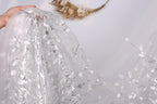 A hand is holding a piece of white fabric with silver leaf patterns, which appears to be a sheer or sheer lace material.