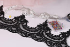 a close-up view of a black lace trim, a measuring tape, a pair of scissors, and a container of colorful pins arranged on a white surface.