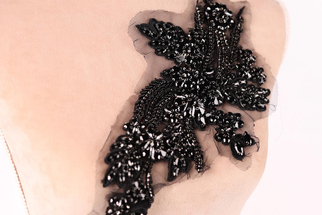 A close-up image of a black, intricate embroidered piece of fabric or material, featuring a floral or leaf-like design with numerous small black beads or sequins attached to it.