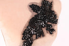 A close-up image of a black, intricate embroidered piece of fabric or material, featuring a floral or leaf-like design with numerous small black beads or sequins attached to it.