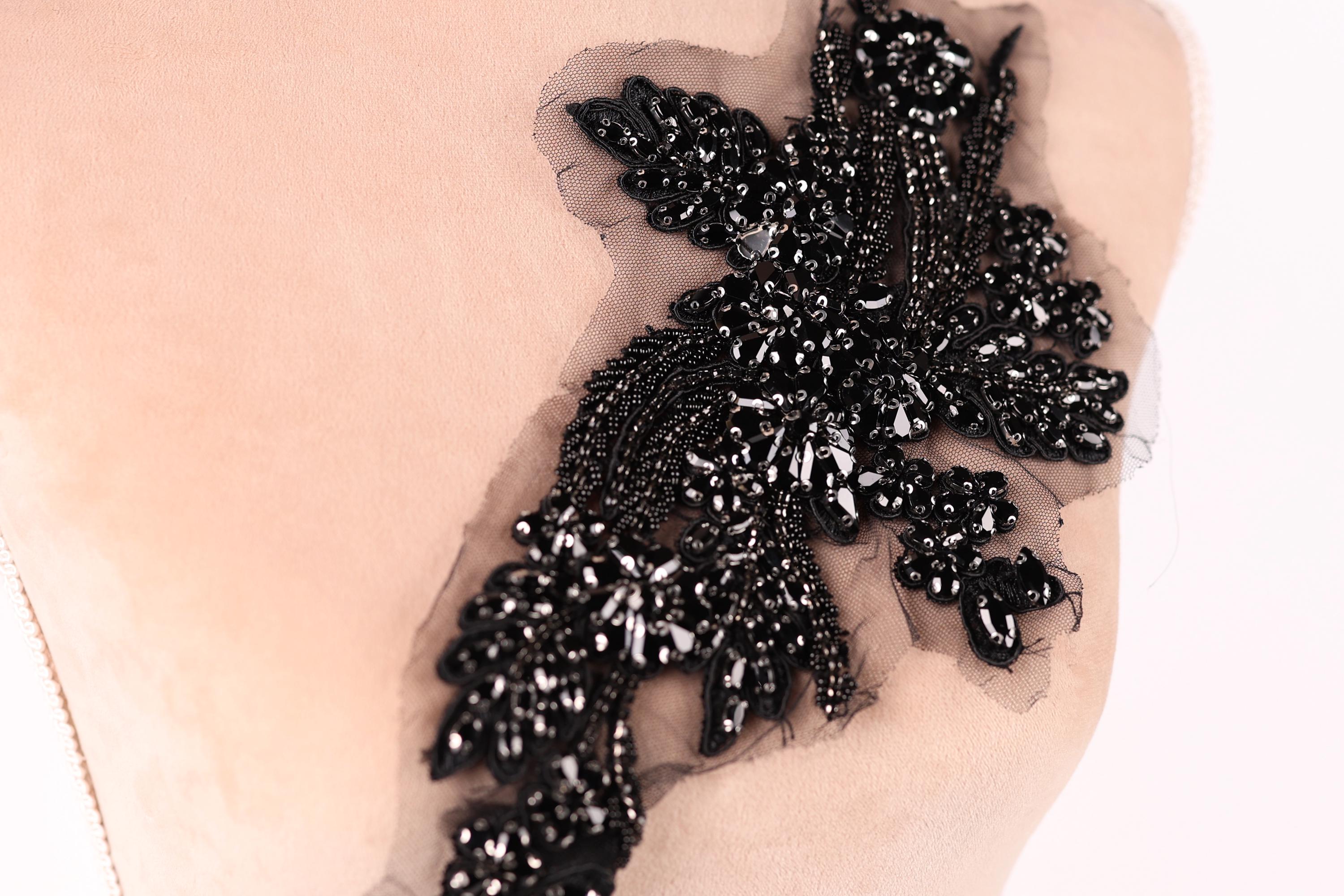 A close-up image of a black, intricate embroidered piece of fabric or material, featuring a floral or leaf-like design with numerous small black beads or sequins attached to it.
