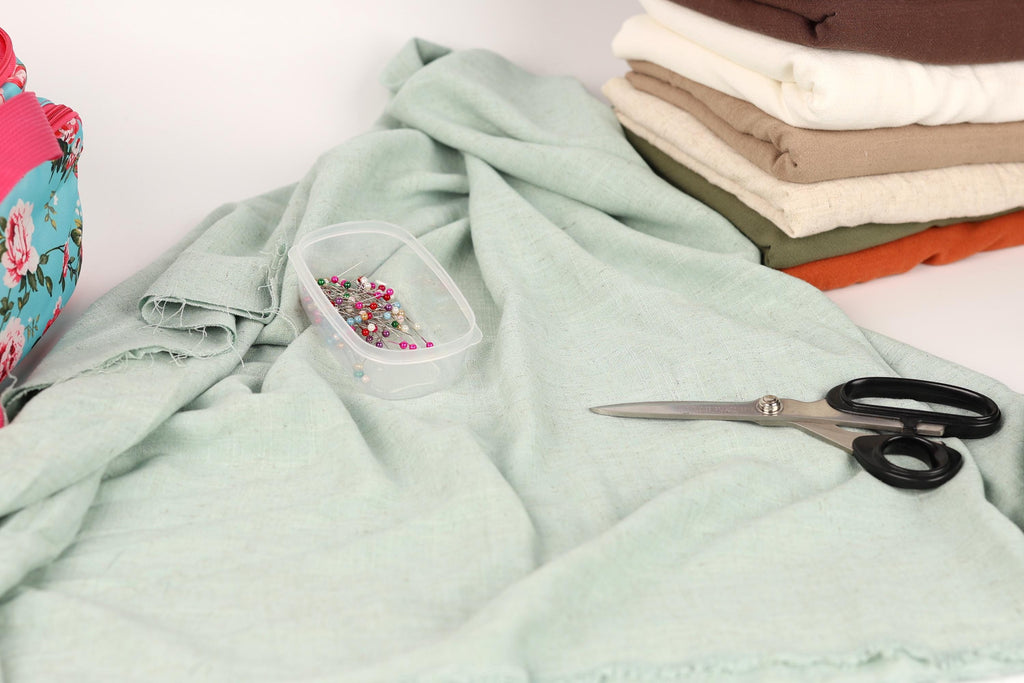 a pile of fabric, a pair of scissors, and a container of pins on a light green cloth.