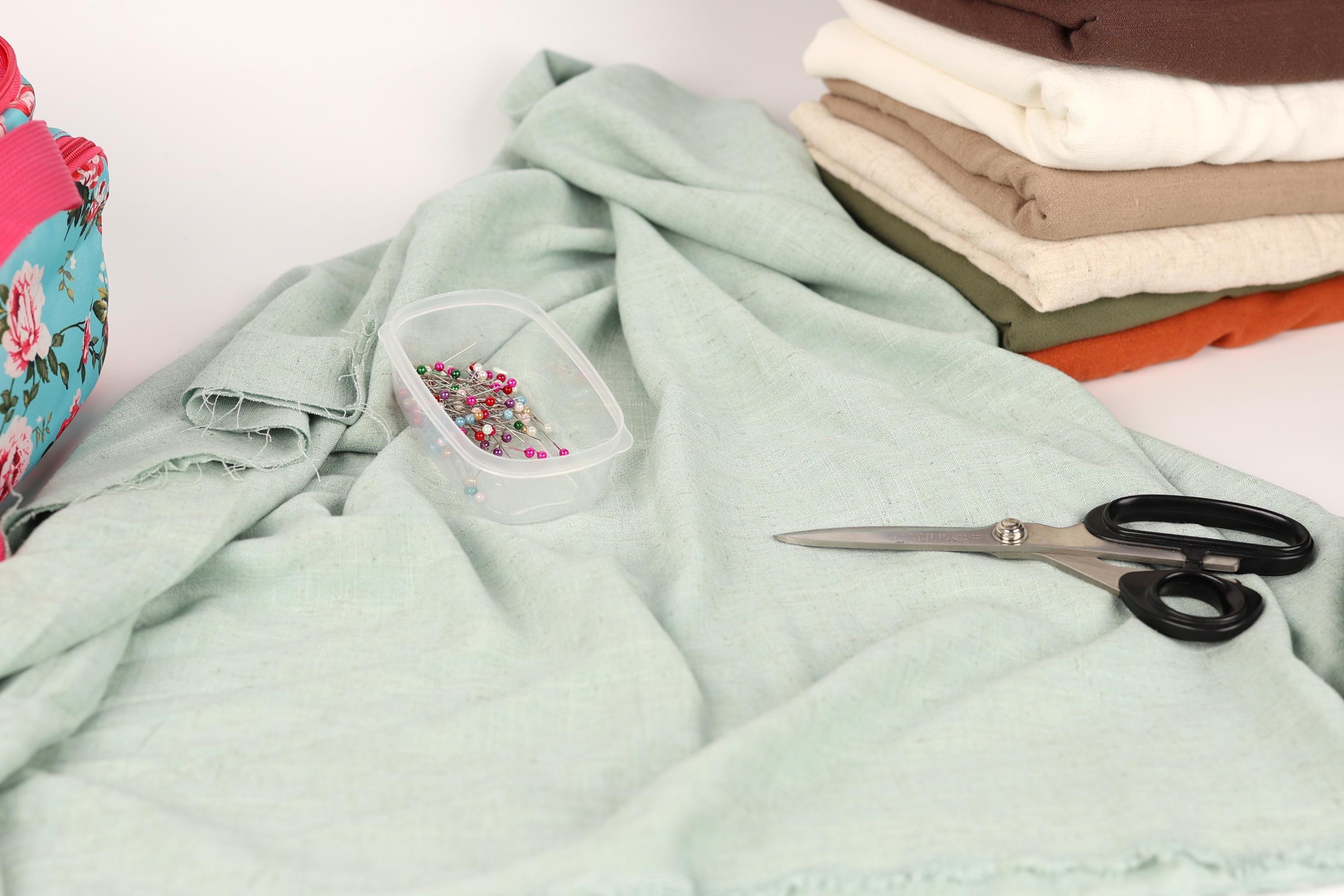 a pile of fabric, a pair of scissors, and a container of pins on a light green cloth.