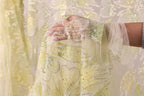 A hand is holding a piece of fabric with a floral pattern and sparkling embellishments, against a background of a white and green floral pattern.