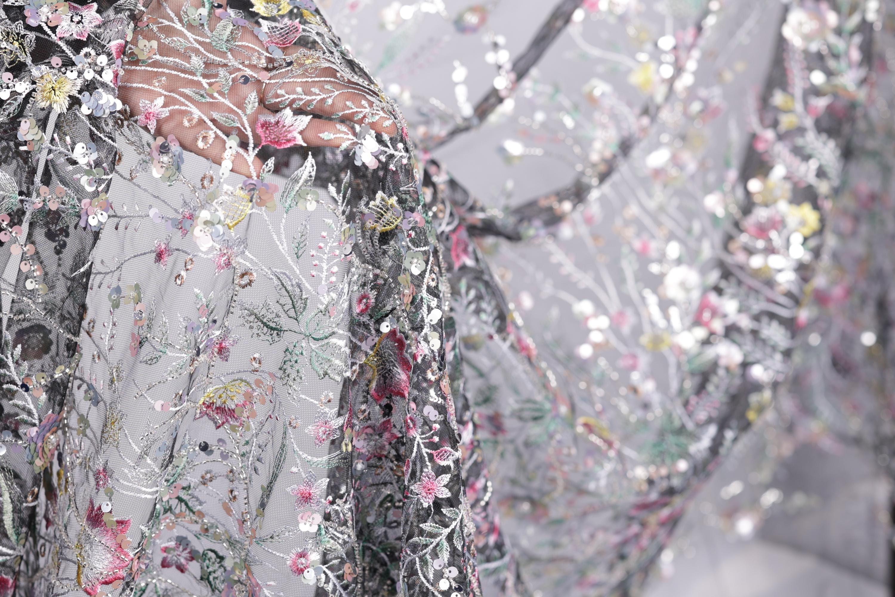 a close-up view of a delicate, intricately embroidered fabric with a floral pattern. The fabric appears to be a sheer material, with a variety of colors and designs, including pink, green, and white flowers. The image is a close-up of the fabric, showcasing the detailed embroidery and the intricate design elements.