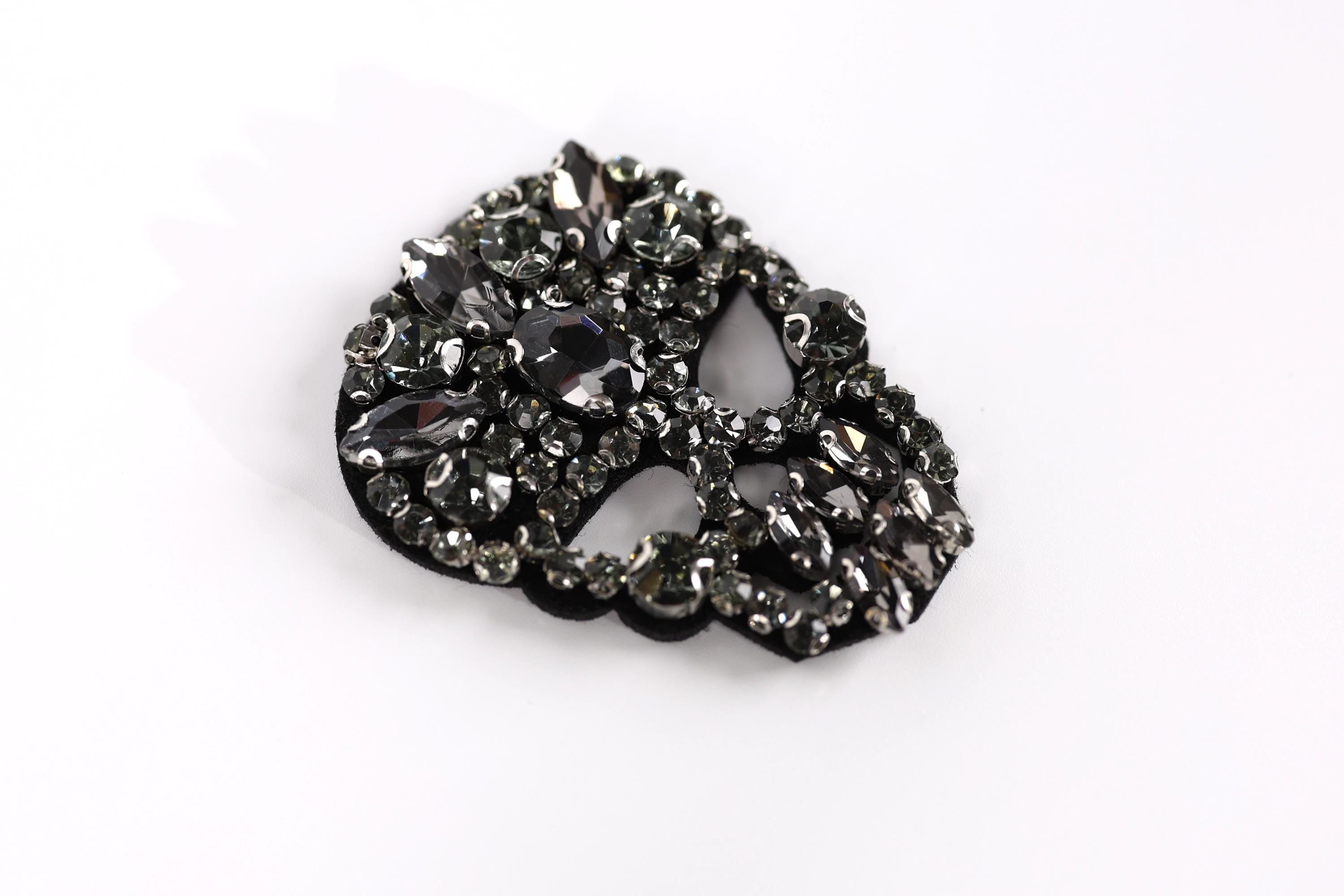 A close-up image of a black and silver rhinestone-encrusted object, possibly a brooch or a piece of jewelry, against a white background.