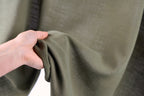 A hand is holding a piece of olive green fabric, possibly a garment or textile, against a blurred background.