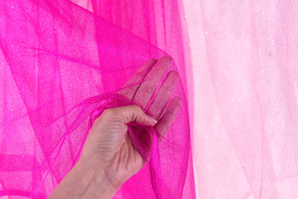 A hand is holding a pink, sparkly fabric up against a pink background.