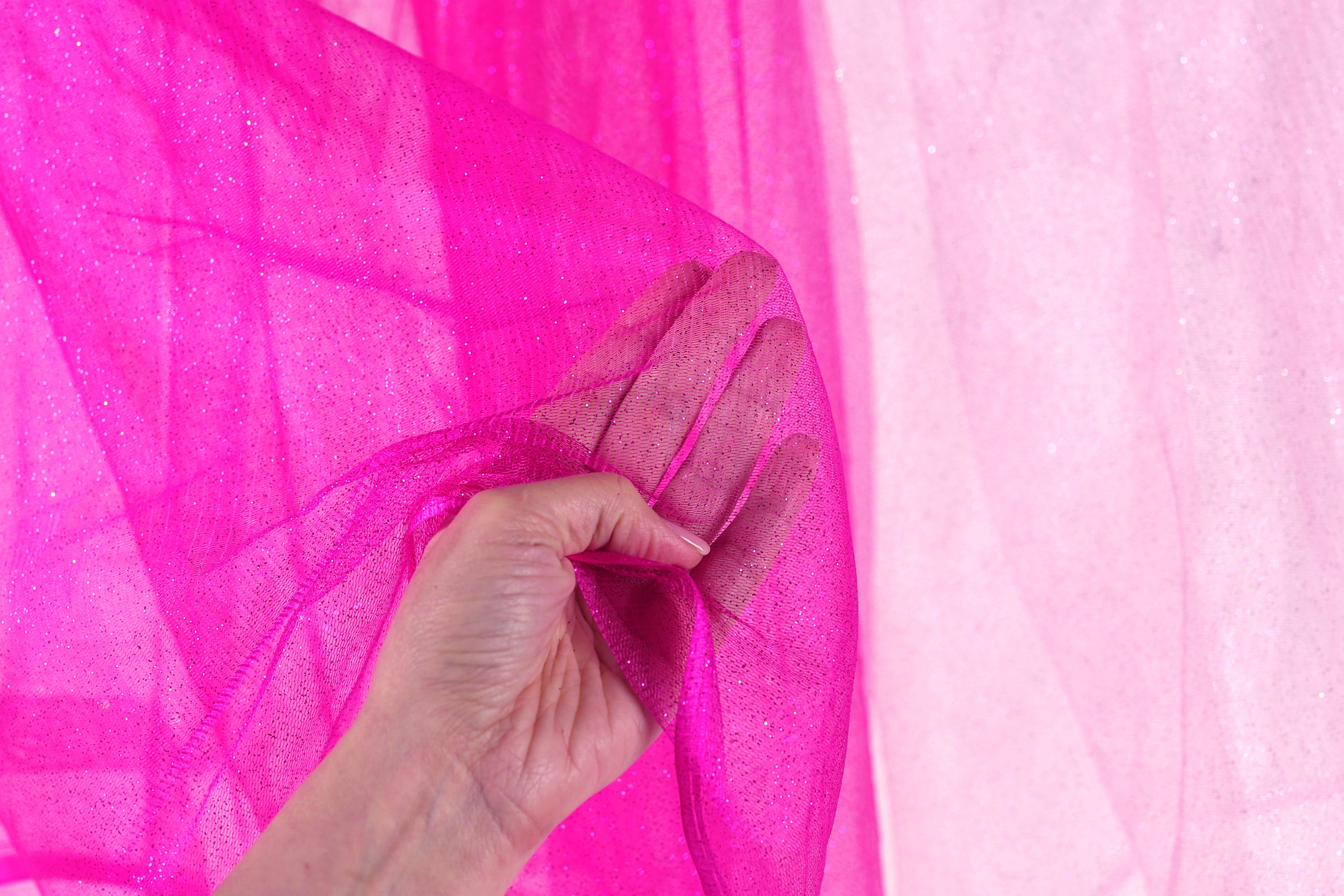 A hand is holding a pink, sparkly fabric up against a pink background.