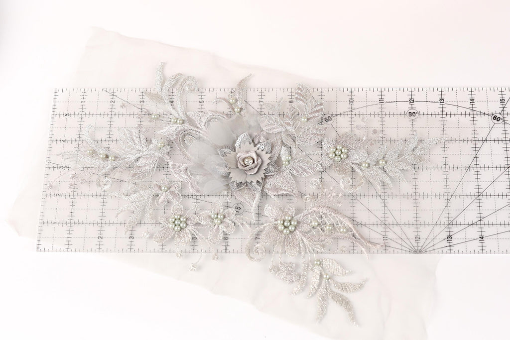 a close-up view of a delicate, intricate piece of embroidery or lace with a floral design. The design includes a large flower surrounded by smaller flowers and leaves, all crafted from a light-colored material. The piece is displayed on a transparent ruler, which is placed on a white surface.