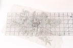 a close-up view of a delicate, intricate piece of embroidery or lace with a floral design. The design includes a large flower surrounded by smaller flowers and leaves, all crafted from a light-colored material. The piece is displayed on a transparent ruler, which is placed on a white surface.