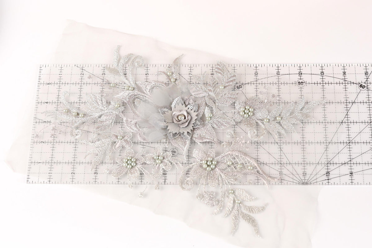 a close-up view of a delicate, intricate piece of embroidery or lace with a floral design. The design includes a large flower surrounded by smaller flowers and leaves, all crafted from a light-colored material. The piece is displayed on a transparent ruler, which is placed on a white surface.