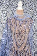 a mannequin wearing a sheer, sequined dress with a blue and silver pattern. The dress has a high neckline and a long, flowing cape-like design. The background is a gray and white patterned fabric.