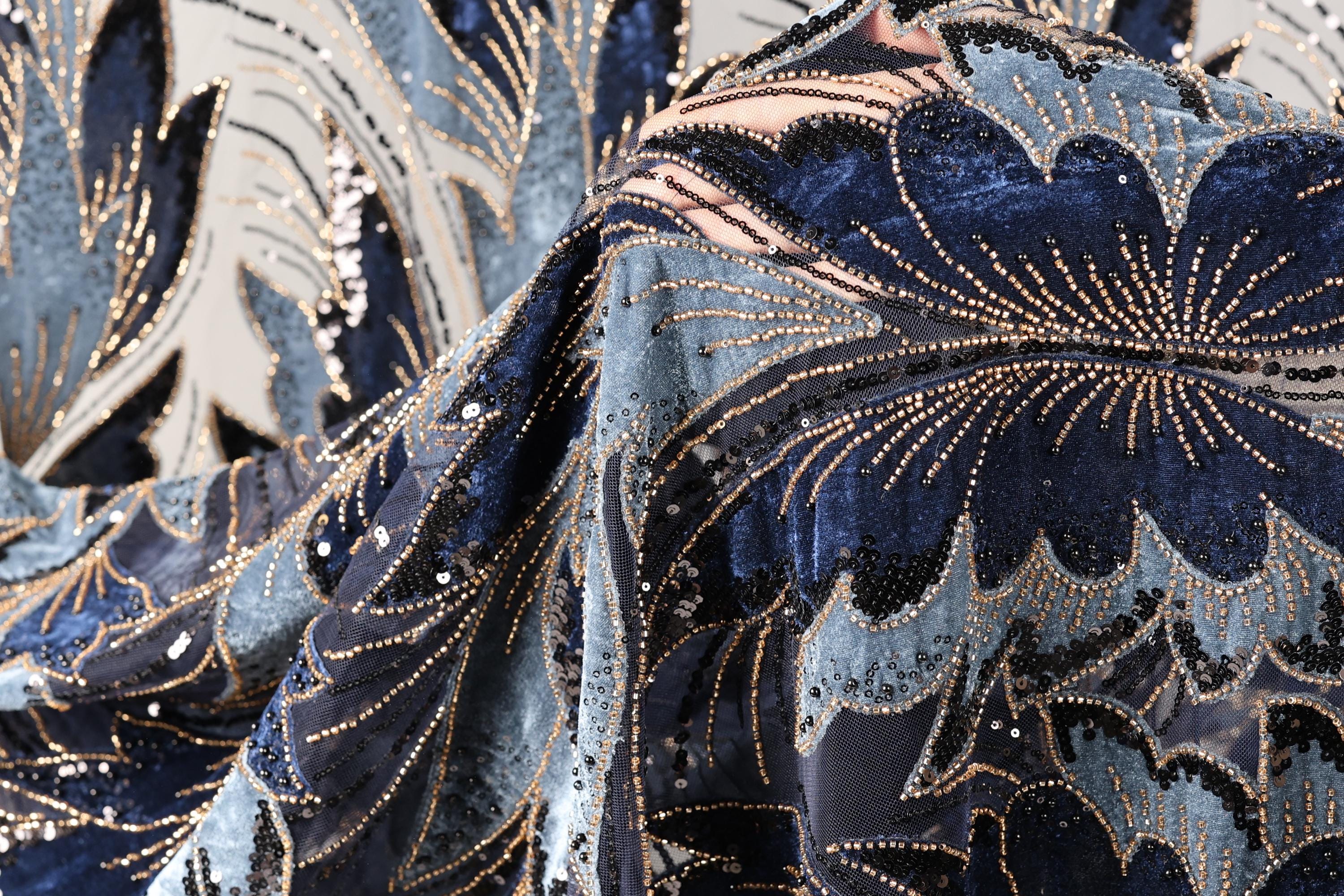 a close-up view of a fabric with a floral pattern, featuring a mix of blue, black, and gold colors. The fabric appears to be made of a shiny material, possibly sequin or beads, and has a textured appearance.