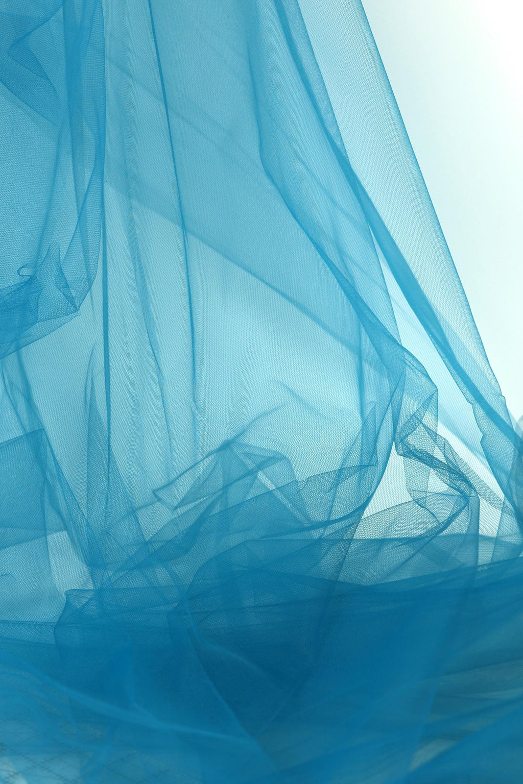 The image is a close-up of a blue fabric, possibly a sheer or sheer-like material, with a soft, flowing texture. The fabric appears to be crumpled or wrinkled, giving it a sense of movement and softness. The color of the fabric is a bright blue, which stands out against the white background. The image does not contain any discernible text or countable objects, and the overall composition is simple and minimalistic.