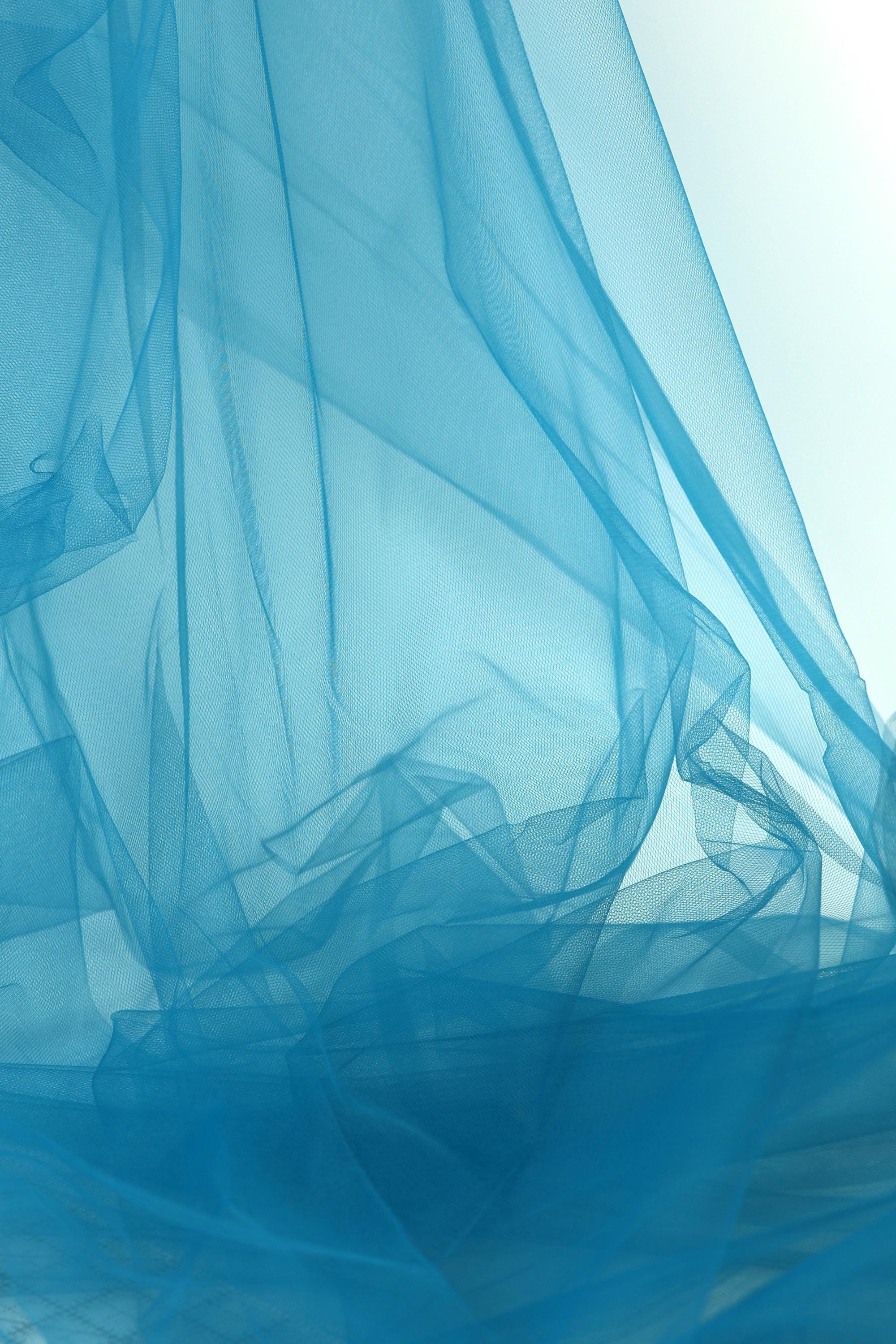 The image is a close-up of a blue fabric, possibly a sheer or sheer-like material, with a soft, flowing texture. The fabric appears to be crumpled or wrinkled, giving it a sense of movement and softness. The color of the fabric is a bright blue, which stands out against the white background. The image does not contain any discernible text or countable objects, and the overall composition is simple and minimalistic.