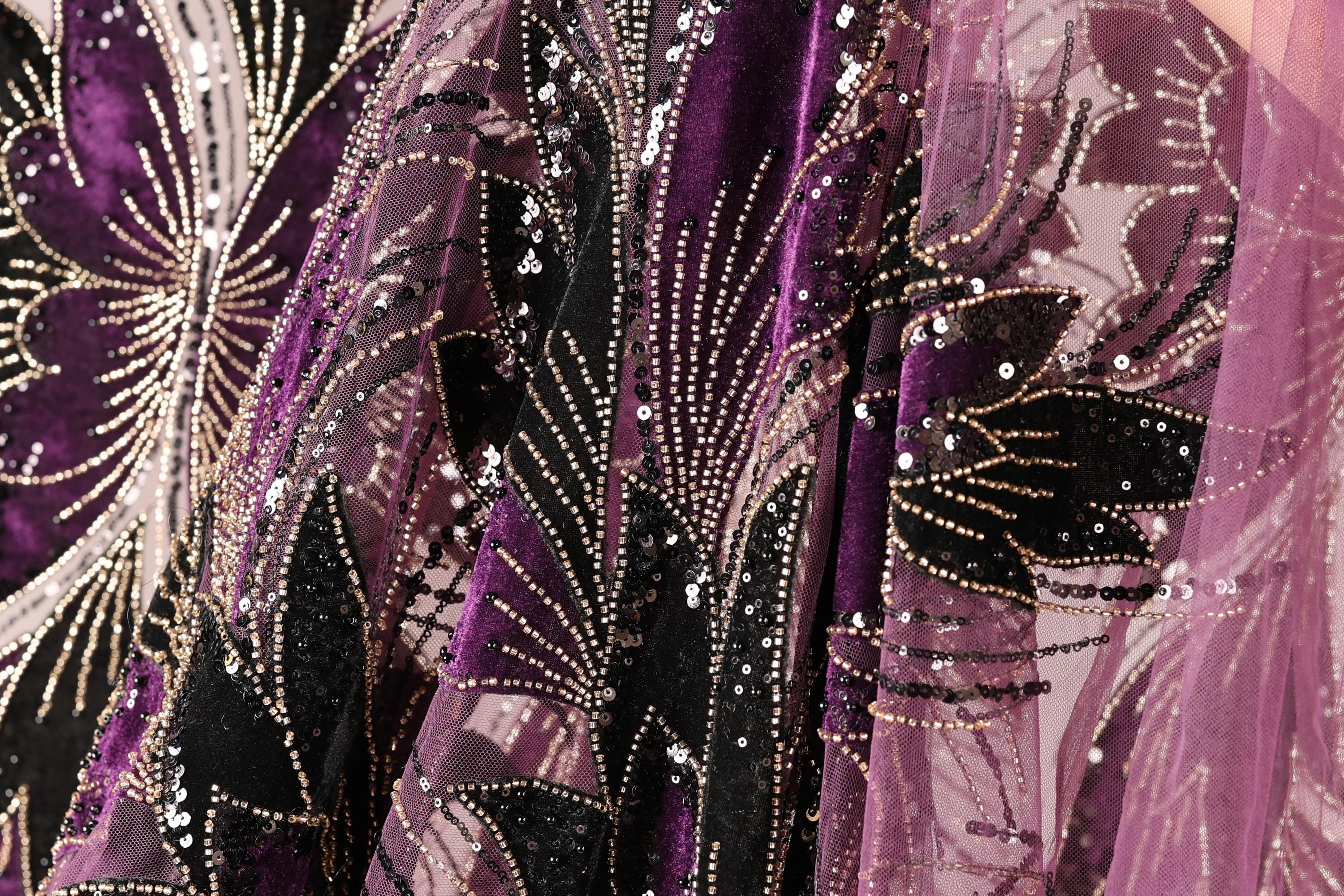 a close-up view of a fabric with a floral pattern, featuring a mix of purple and black colors. The fabric appears to be made of a sheer material, with sequins and beads adding a decorative touch.