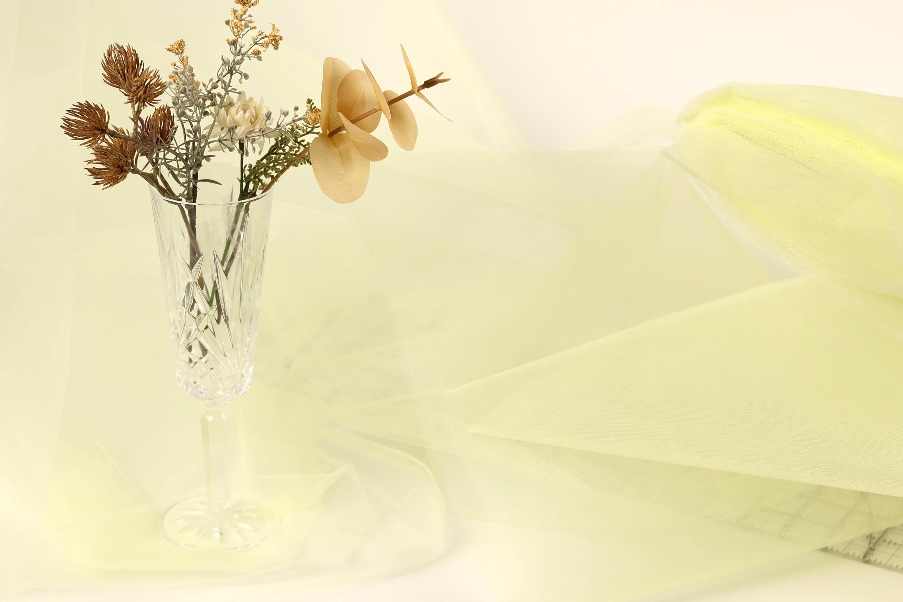 A clear glass vase filled with dried flowers and branches sits on a white surface, with a yellow fabric or material in the background.