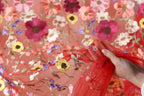 A hand is holding a red fabric with a floral pattern, featuring a variety of colorful flowers such as pink, yellow, and purple.