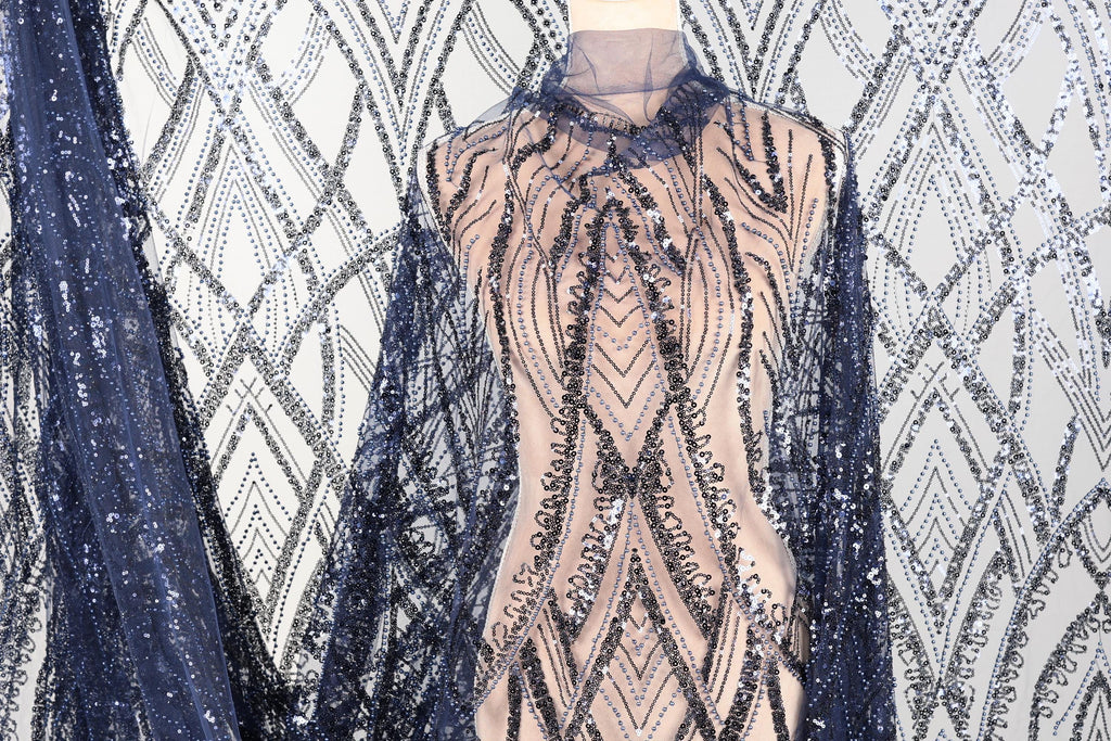 a mannequin wearing a dress with a patterned fabric and a sheer overlay. The dress has a beige color and is adorned with sequins and a lace-like design. The mannequin is positioned in front of a background with a similar patterned fabric, creating a visually cohesive display.