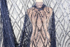 a mannequin wearing a dress with a patterned fabric and a sheer overlay. The dress has a beige color and is adorned with sequins and a lace-like design. The mannequin is positioned in front of a background with a similar patterned fabric, creating a visually cohesive display.