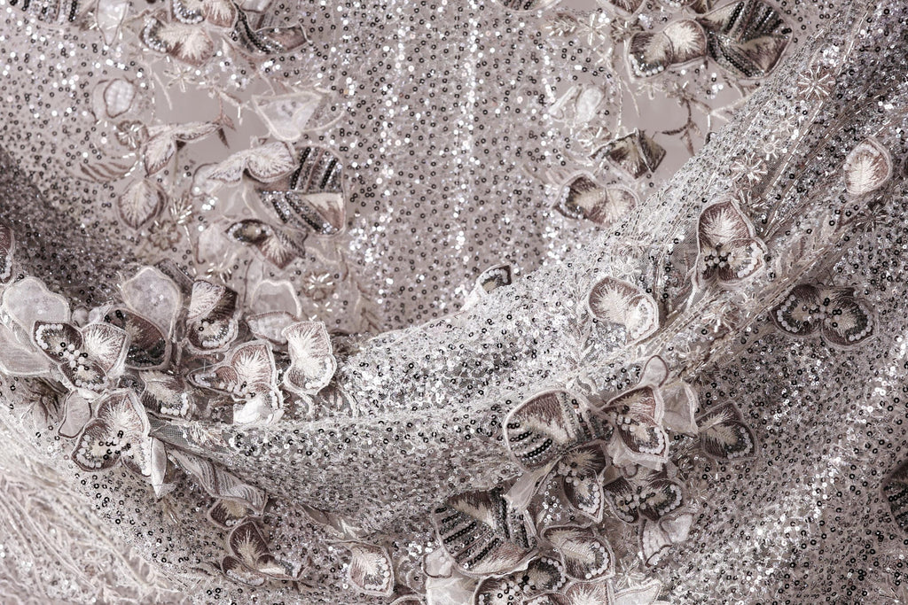 The image is a close-up of a fabric with a silver sequin pattern, featuring a variety of small, intricate designs.