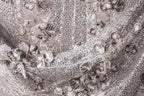 The image is a close-up of a fabric with a silver sequin pattern, featuring a variety of small, intricate designs.
