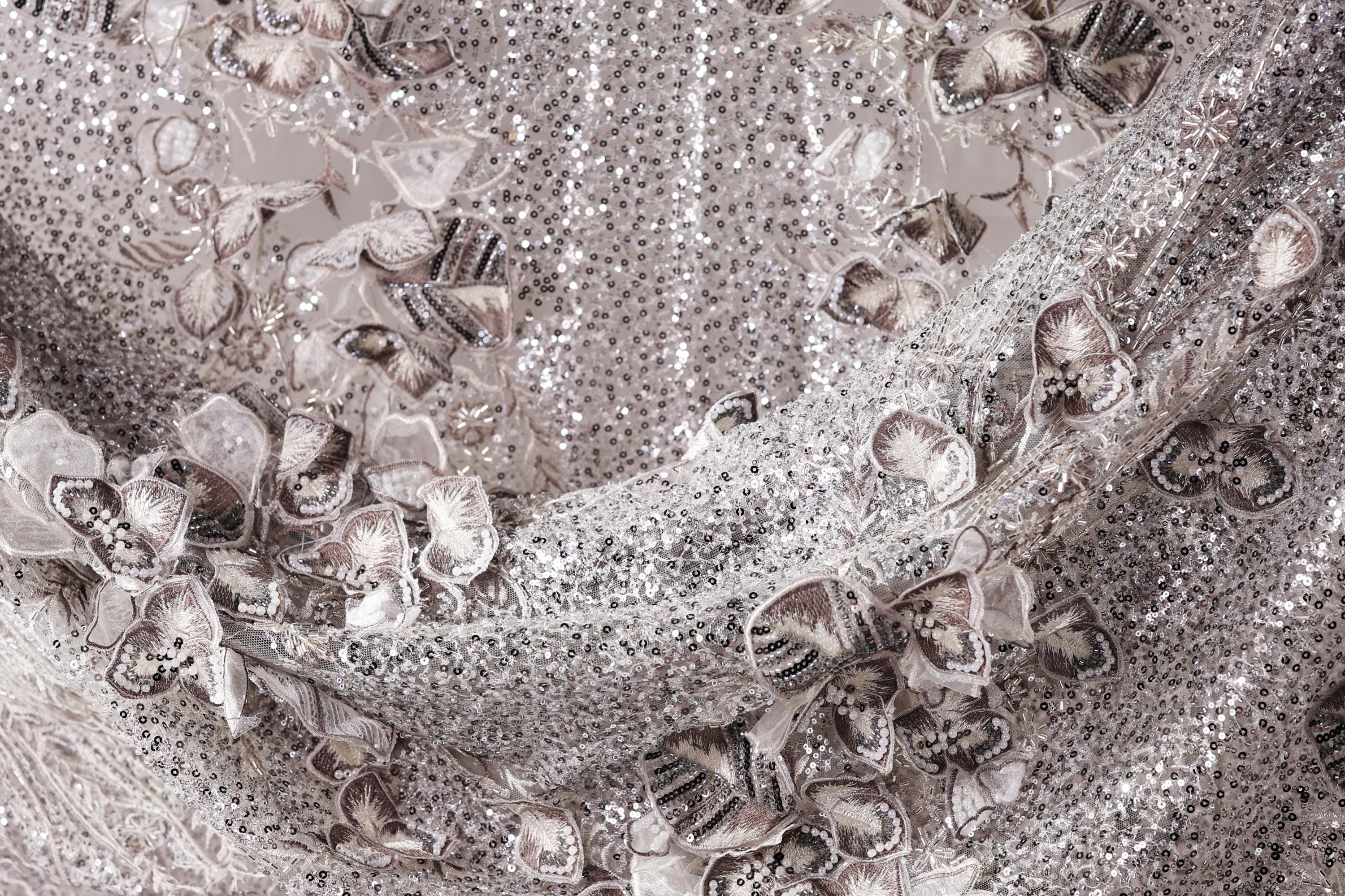 The image is a close-up of a fabric with a silver sequin pattern, featuring a variety of small, intricate designs.