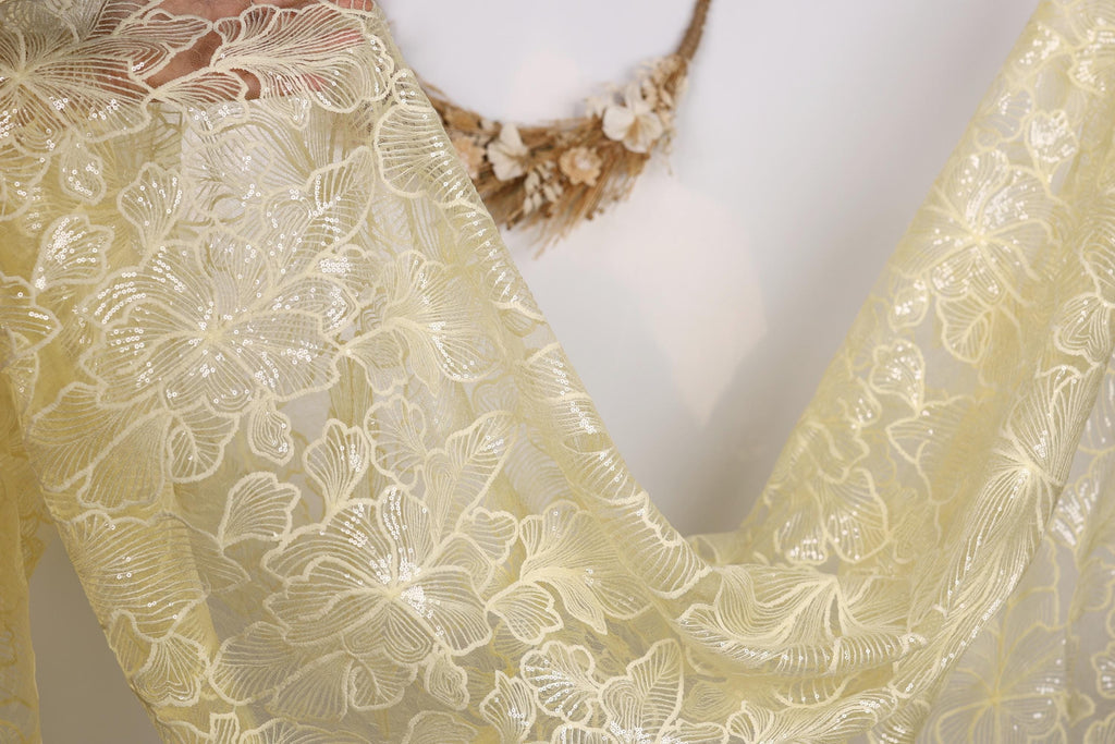 a close-up view of a delicate, yellow lace fabric with a floral pattern. The fabric appears to be made of sheer material, giving it a soft and delicate appearance. The background is a plain white wall, which allows the focus to remain on the intricate details of the lace fabric.