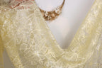 a close-up view of a delicate, yellow lace fabric with a floral pattern. The fabric appears to be made of sheer material, giving it a soft and delicate appearance. The background is a plain white wall, which allows the focus to remain on the intricate details of the lace fabric.