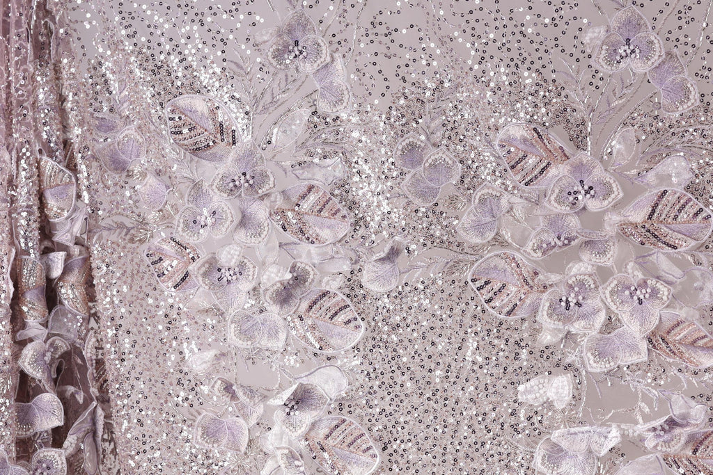 a close-up view of a fabric with a floral pattern, including purple and white flowers and sequins. The fabric appears to be made of a shiny, glittery material, giving it a sparkling and elegant appearance.