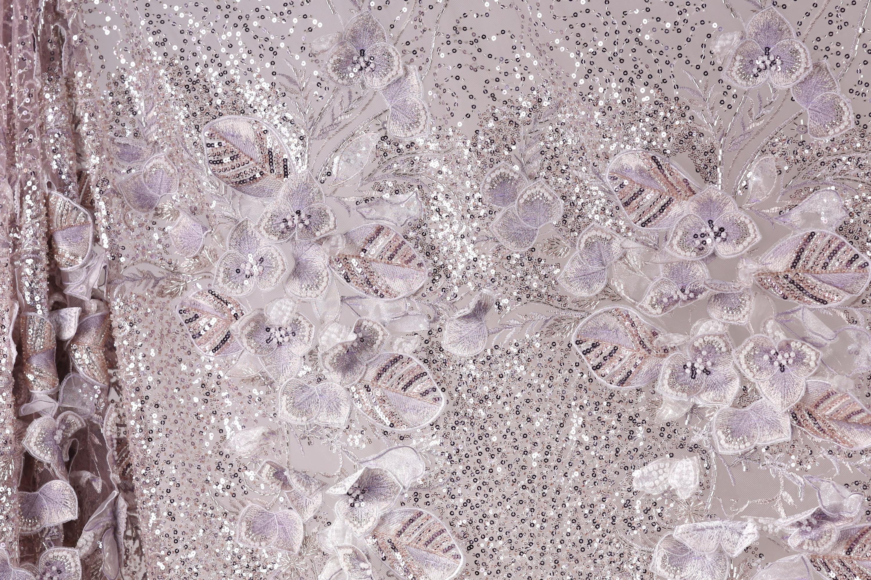 a close-up view of a fabric with a floral pattern, including purple and white flowers and sequins. The fabric appears to be made of a shiny, glittery material, giving it a sparkling and elegant appearance.