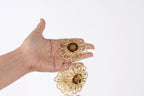 A hand holding two gold flower-shaped earrings against a white background.