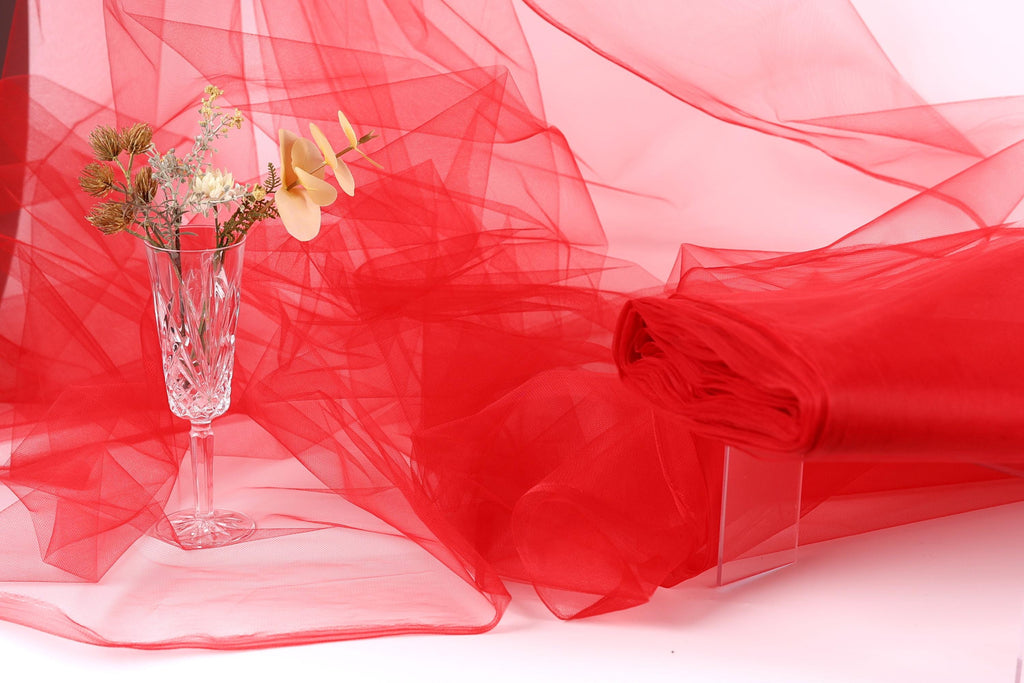 a clear glass vase filled with flowers, placed on a white surface next to a red fabric or sheer material.