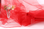 a clear glass vase filled with flowers, placed on a white surface next to a red fabric or sheer material.