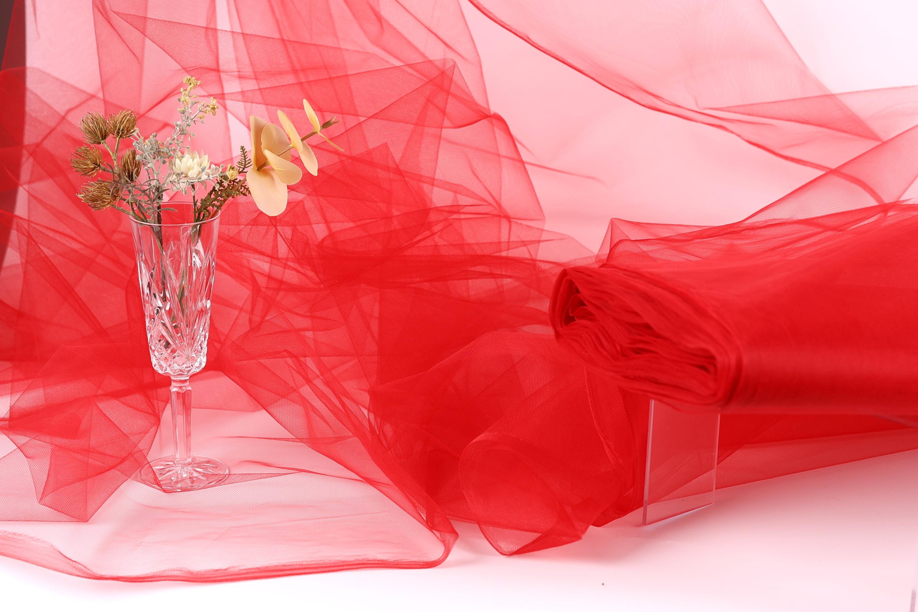 a clear glass vase filled with flowers, placed on a white surface next to a red fabric or sheer material.