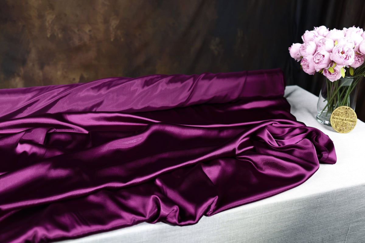 a purple satin fabric draped over a white table, with a vase of pink flowers placed next to it.