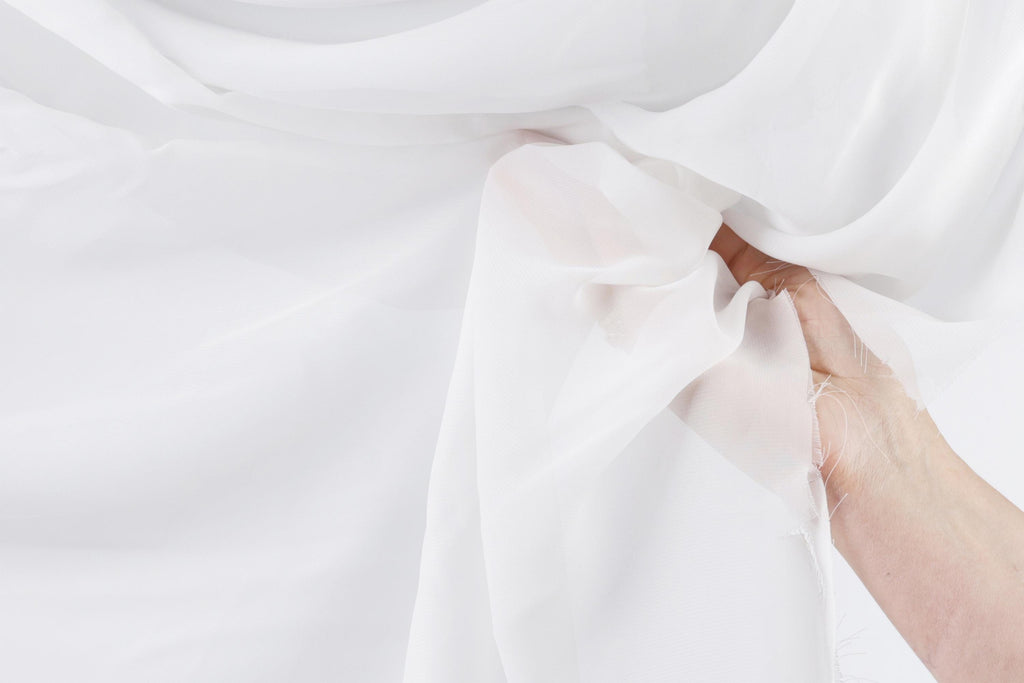 A hand is holding a white fabric, possibly a sheet or a cloth, against a plain white background.