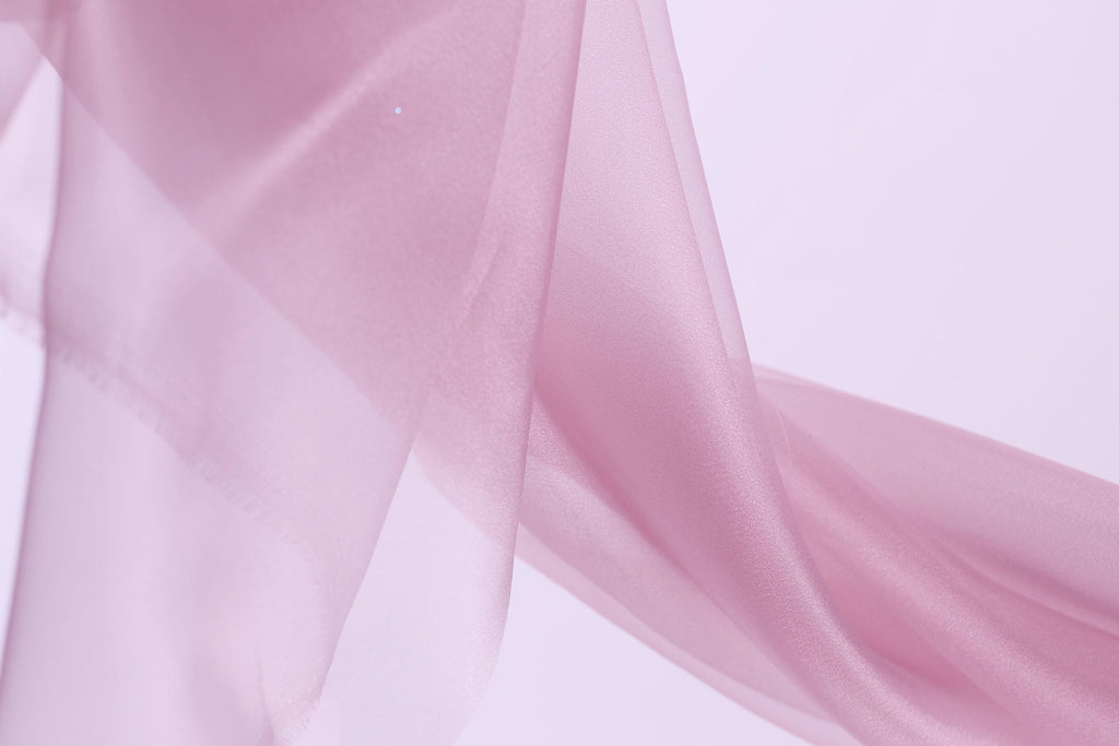 A close-up view of a soft, flowing pink fabric, possibly silk or chiffon, against a plain white background.